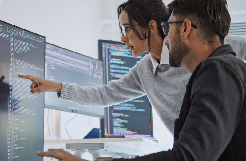 software-engineers-point-at-code-while-working-on-desktop-computer-is-2156387160 Software engineers collaborating on coding projects at home office using computers.